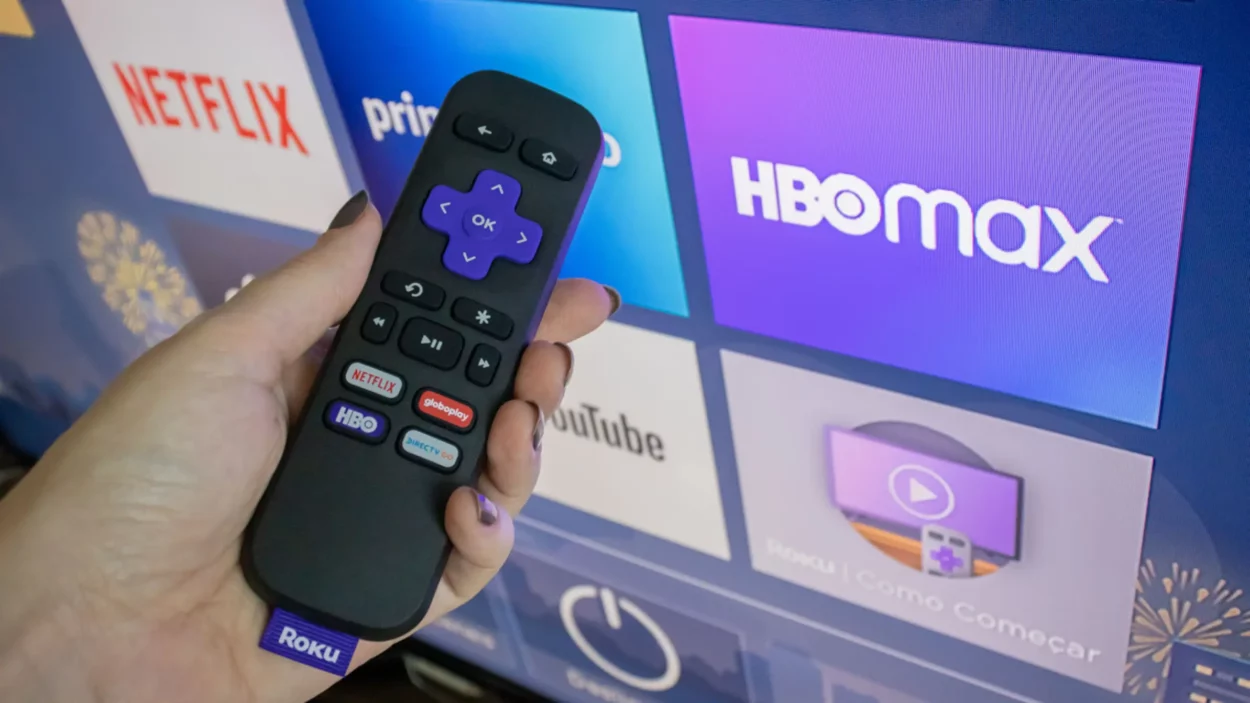 A hand holding a TCL Roku TV remote in front of TCL Roku TV