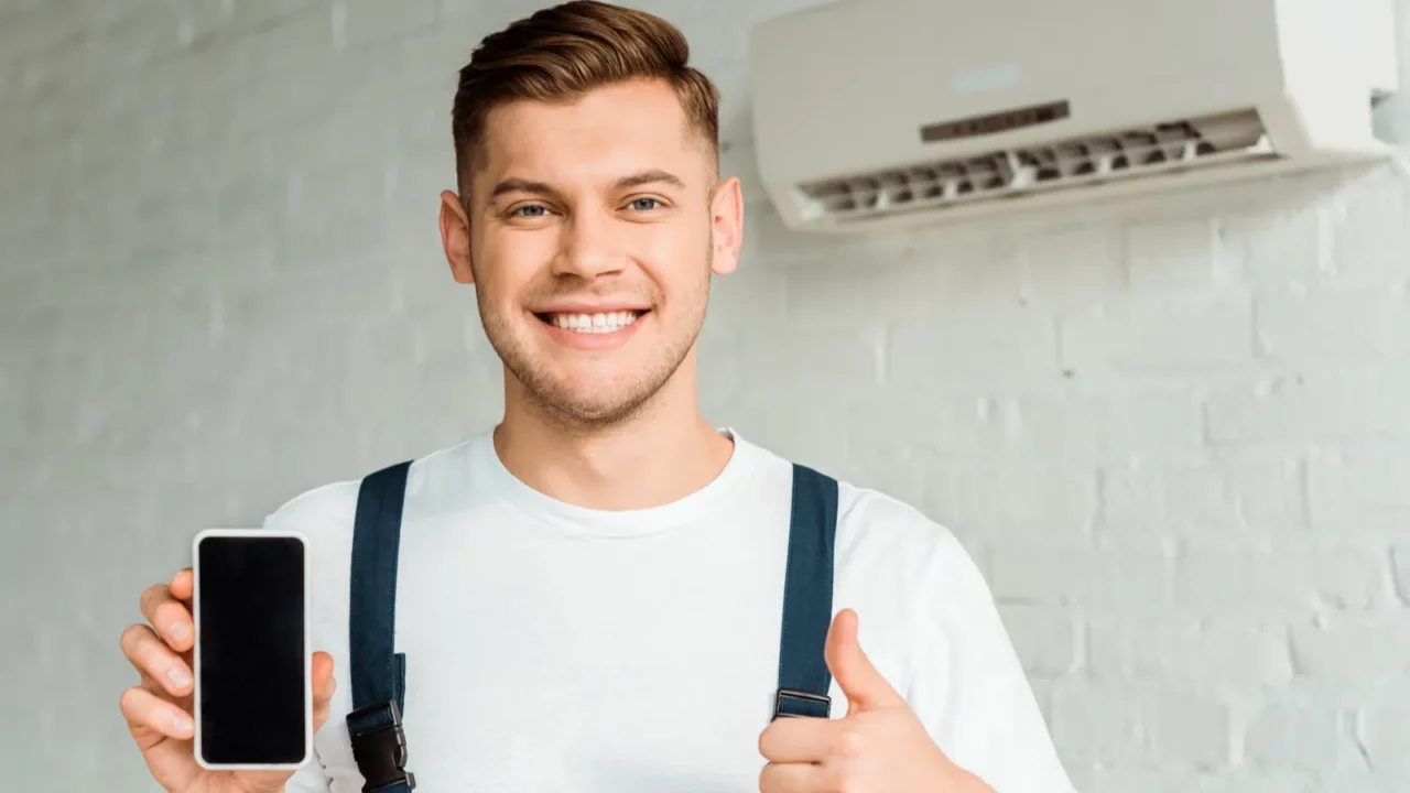 cheerful installer showing thumb up while holding smartphone with blank