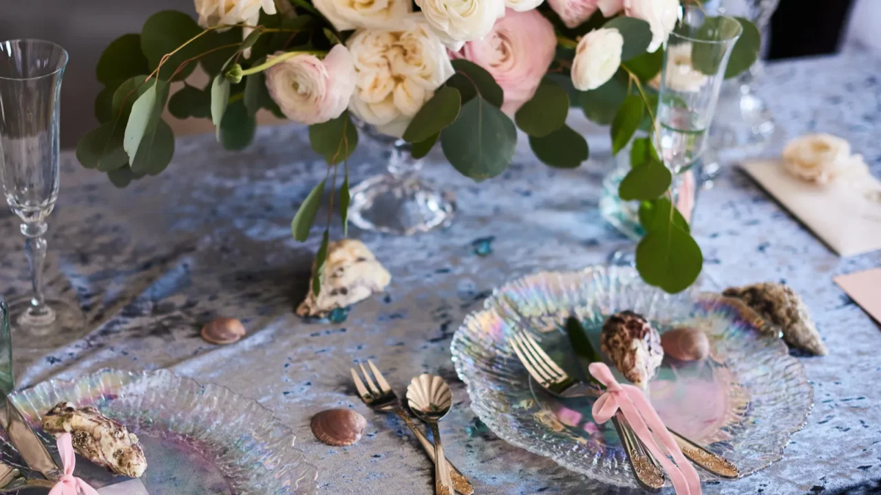 Elegant table setting with iridescent plates, pink-ribboned cutlery, seashells, and a floral centerpiece of white and pink roses.