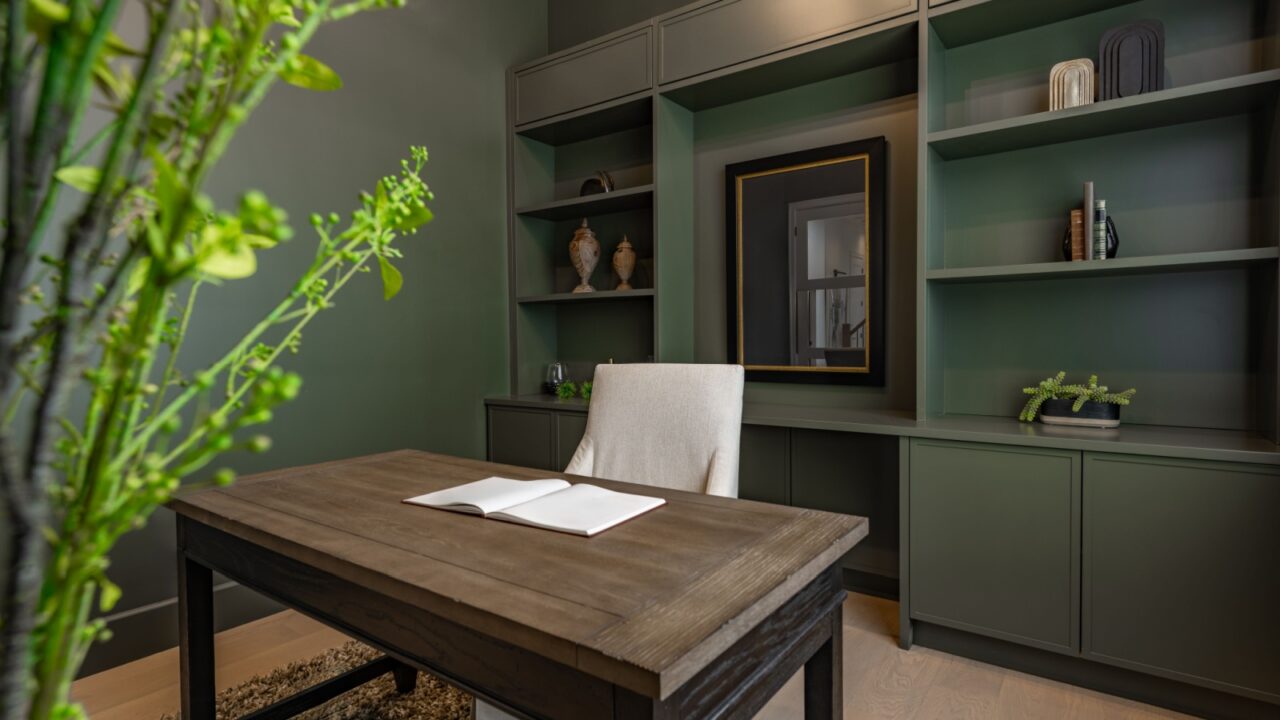 Home office study den interior with built in shelving dark wooden desk upholstered chair tall houseplant in foreground blurred deep green painted walls and shelves cupboards elegant masculine room