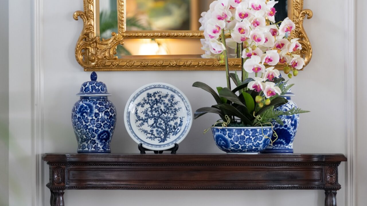 Blue paint porcelain collection with Orchid vase on a dark wooden table with golden mirror on white wall.