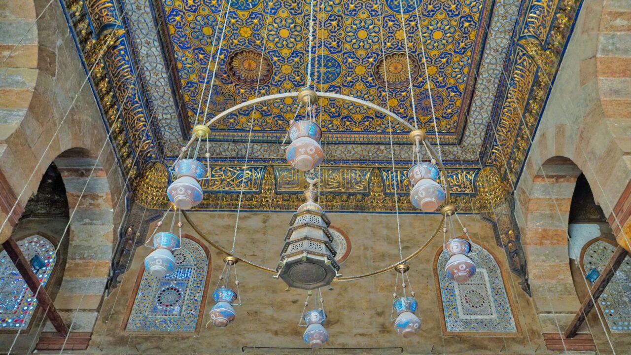 Ornate ceiling with intricate blue and gold patterns, Arabic calligraphy, and hanging lamps in a historical building.