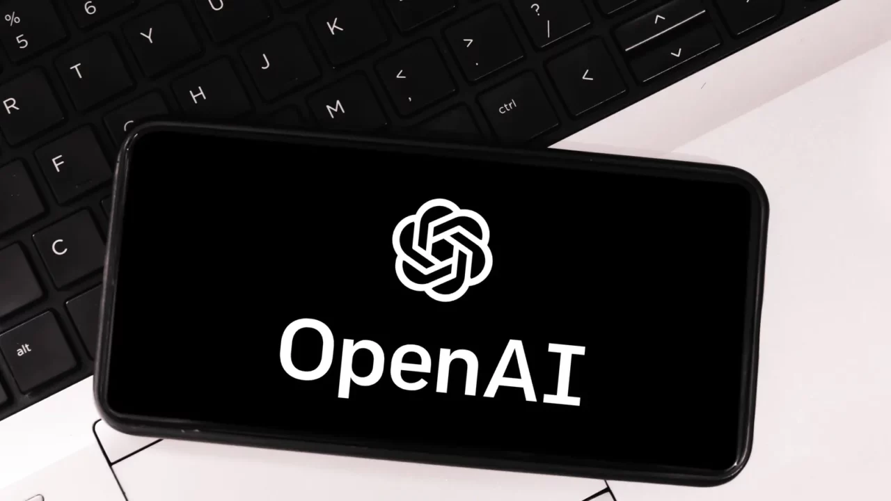 OpenAI logo on mobile screen with laptop in the background.